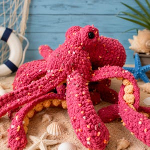 Peut inclure: Une peluche pieuvre fabriquée en fil magenta épais avec une texture mouchetée de taches blanches, jaunes et orange. Le dessous des huit tentacules présente une couleur beige clair contrastante et la tête possède deux yeux noirs.