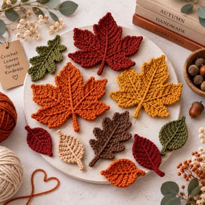 May include: A collection of crocheted leaves in various shapes and autumn colors, including deep red, orange, yellow, brown, beige, and green, arranged on a round white plate. The set includes maple and oak leaf designs with visible yarn texture.