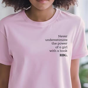 May include: A light pink t-shirt with the text "Never underestimate the power of a girl with a book. RBG." printed in black. The shirt has short sleeves and a crew neck.