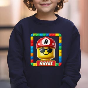 May include: A navy blue long-sleeve sweatshirt featuring a central graphic of a yellow toy figure head wearing a red cap with the letter A and black sunglasses. The graphic is framed by colorful building blocks and includes the name ARIEL.