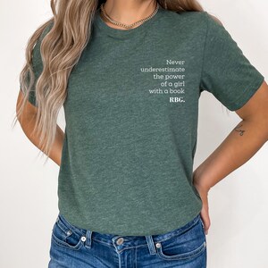 May include: Green t-shirt with the text "Never underestimate the power of a girl with a book. RBC." The shirt is a solid color and has a crew neck. The text is in a white, sans-serif font.