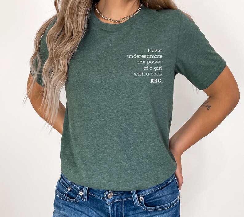 May include: Green t-shirt with the text "Never underestimate the power of a girl with a book. RBC." The shirt is a solid color and has a crew neck. The text is in a white, sans-serif font.