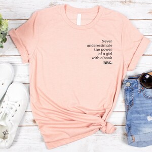 May include: A peach-colored t-shirt with the text "Never underestimate the power of a girl with a book. RBG." is displayed with denim shorts, white sneakers, and sunglasses. The shirt is folded, and the items are arranged on a light wood surface.