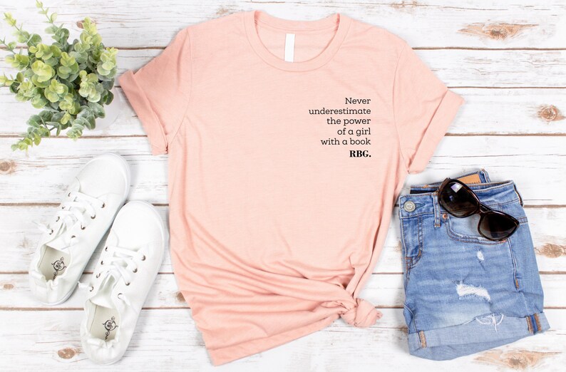 May include: A peach-colored t-shirt with the text "Never underestimate the power of a girl with a book. RBG." is displayed with denim shorts, white sneakers, and sunglasses. The shirt is folded, and the items are arranged on a light wood surface.