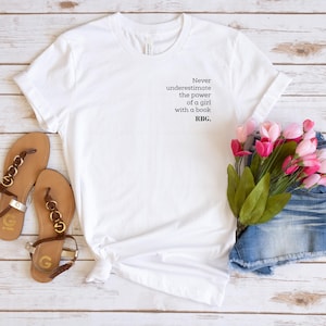 May include: White t-shirt with the text "Never underestimate the power of a girl with a book. RBG." Brown sandals with gold accents and a pair of denim shorts are next to the shirt. Pink flowers complete the scene.
