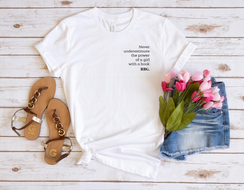 May include: White t-shirt with the text "Never underestimate the power of a girl with a book. RBG." Brown sandals with gold accents and a pair of denim shorts are next to the shirt. Pink flowers complete the scene.
