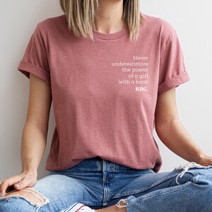 May include: A dusty rose t-shirt with rolled sleeves. The shirt features the text "Never underestimate the power of a girl with a book. RBG." in white. The person is wearing blue jeans with ripped knees.