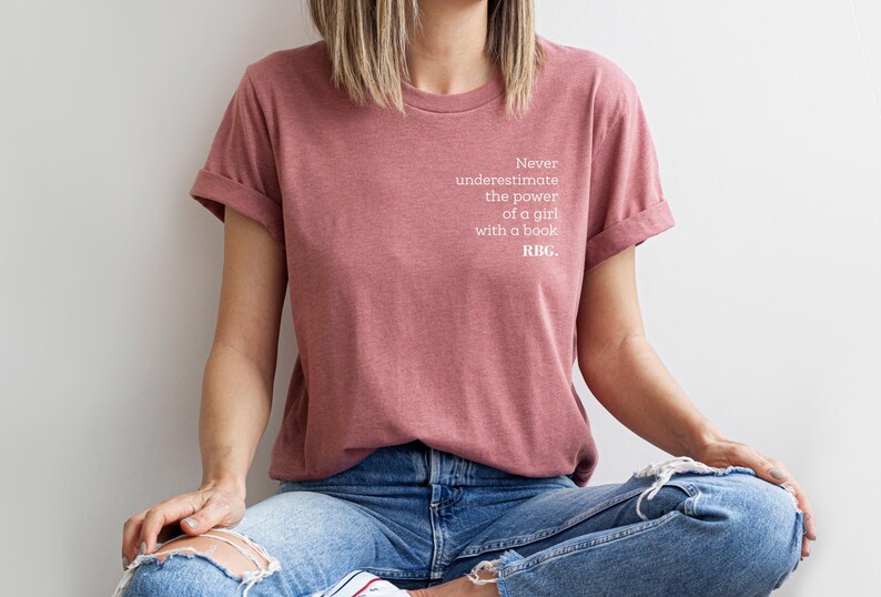May include: A dusty rose t-shirt with rolled sleeves. The shirt features the text "Never underestimate the power of a girl with a book. RBG." in white. The person is wearing blue jeans with ripped knees.