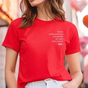 May include: A bright red t-shirt with the text "Never underestimate the power of a girl with a book. RBG." in white. The shirt is short-sleeved and made of a smooth fabric. The background includes blurred lights and heart-shaped balloons.