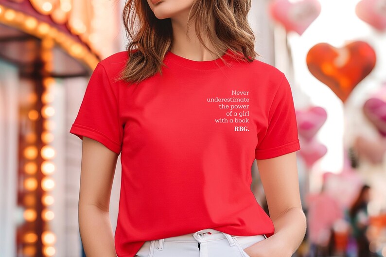 May include: A bright red t-shirt with the text "Never underestimate the power of a girl with a book. RBG." in white. The shirt is short-sleeved and made of a smooth fabric. The background includes blurred lights and heart-shaped balloons.
