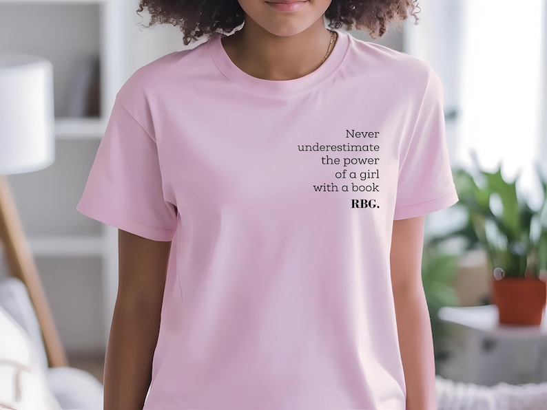 May include: A light pink t-shirt with the text "Never underestimate the power of a girl with a book. RBG." printed in black. The shirt has short sleeves and a crew neck.