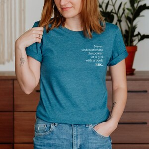 May include: Teal-colored t-shirt with the quote "Never underestimate the power of a girl with a book. RBG." printed in white. The shirt is made of a soft material and is suitable for casual wear.