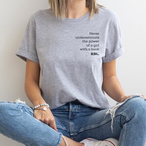 May include: A light gray t-shirt with rolled-up sleeves featuring the quote, "Never underestimate the power of a girl with a book. RBG." The shirt is paired with blue jeans. The person is sitting with their legs crossed.