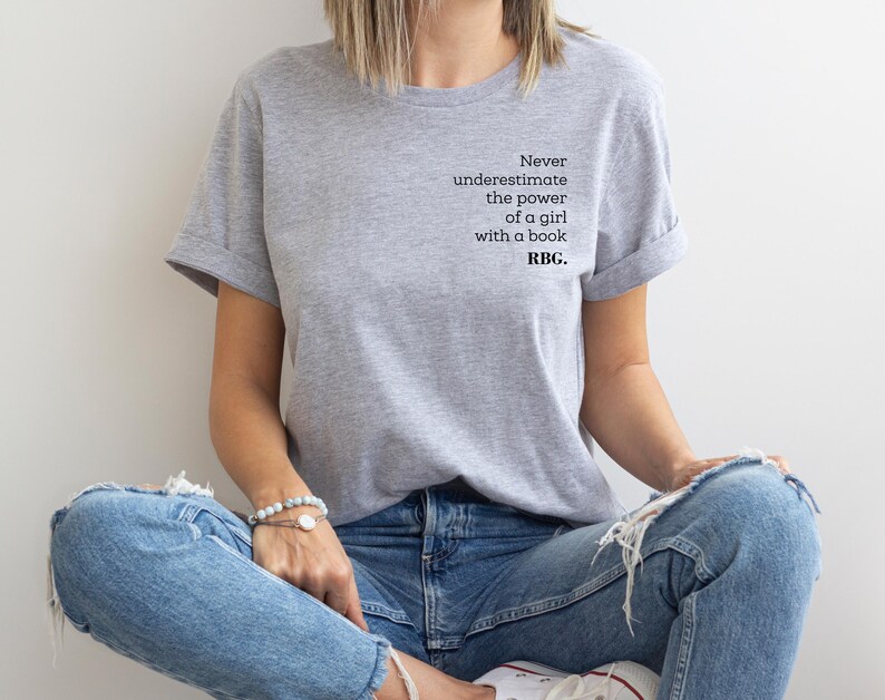 May include: A light gray t-shirt with rolled-up sleeves featuring the quote, "Never underestimate the power of a girl with a book. RBG." The shirt is paired with blue jeans. The person is sitting with their legs crossed.