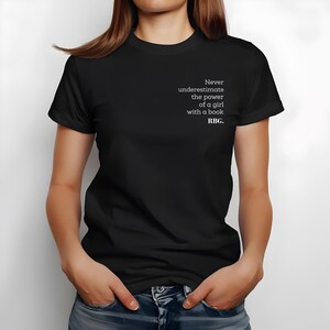May include: Black t-shirt with the text "Never underestimate the power of a girl with a book. RBG." The text is in white, and the shirt is being worn by a person. The person is wearing blue jeans.