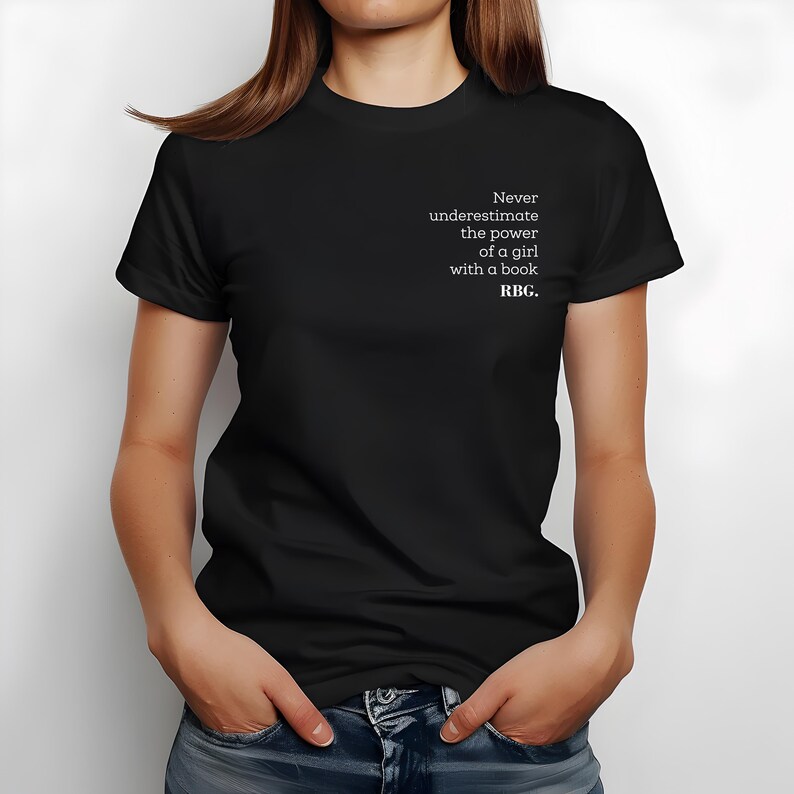 May include: Black t-shirt with the text "Never underestimate the power of a girl with a book. RBG." The text is in white, and the shirt is being worn by a person. The person is wearing blue jeans.