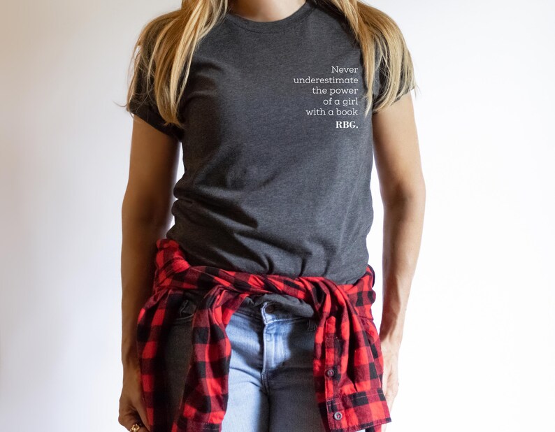 May include: Dark gray t-shirt with the text "Never underestimate the power of a girl with a book. RBG." A red and black plaid shirt is tied around the waist. The person is wearing blue jeans.