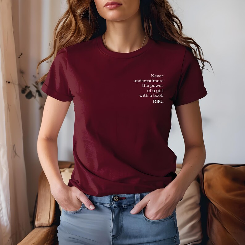 May include: A maroon t-shirt with the text "Never underestimate the power of a girl with a book. RBG." in white. The shirt is a crew neck style and is paired with blue jeans.