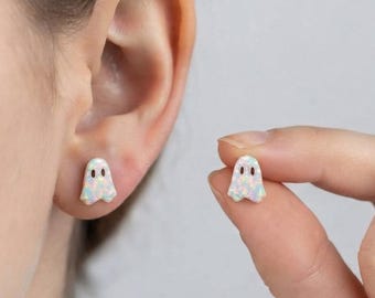 May include: A pair of stud earrings shaped like small ghosts. Each ghost is crafted from a white iridescent material with a subtle color-shifting finish and features two small, dark, circular indentations for eyes. One earring is worn in an earlobe.