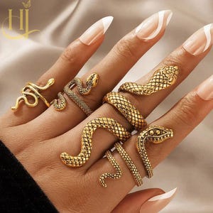 May include: A collection of four gold-toned rings featuring snake designs. The rings display textured, scale-like patterns on the bodies of the snakes, with some designs wrapping around the fingers in coiled shapes and others featuring distinct heads.