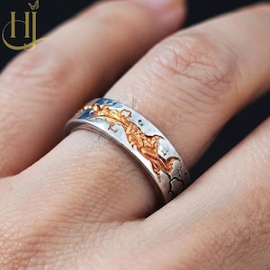May include: A silver-toned band ring featuring a recessed, textured gold-colored inlay that depicts a winding dragon motif. The metal surface has a weathered, oxidized finish with dark etched lines surrounding the central golden dragon design.