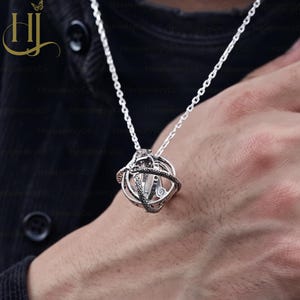 May include: A silver-toned spherical pendant necklace featuring an intricate armillary sphere design with multiple interlocking rings. The metal surface displays detailed engravings and a textured finish, suspended from a matching silver-toned chain.