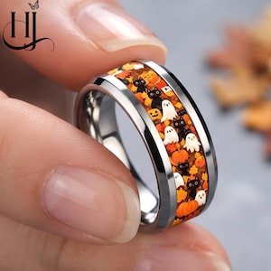 May include: A silver-toned metal ring featuring a central inlay band decorated with a repeating pattern of black cats, white ghosts, orange pumpkins, and autumn leaves in shades of yellow, orange, and brown against a dark background.