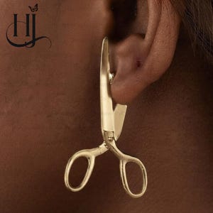 May include: A gold-toned earring shaped like a pair of open scissors. The earring features two oval finger loops at the bottom and two pointed blades extending upward, with the pivot point positioned near the center of the scissor-shaped design.