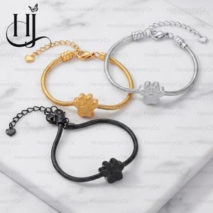 May include: Three snake chain bracelets featuring a central paw print charm. One bracelet is gold-toned, one is silver-toned, and one is black. Each bracelet includes an adjustable chain with a small heart-shaped charm at the end and a lobster clasp.