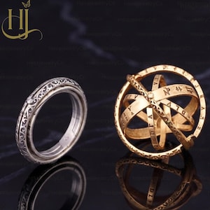 May include: A silver-toned ring with intricate engraved patterns alongside a gold-toned spherical armillary ring. The armillary ring features multiple interlocking bands inscribed with various symbols and letters, designed to unfold into a sphere.