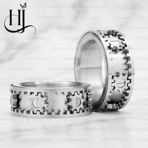 May include: A silver-toned metal ring featuring a rotating outer band with a series of raised gear-shaped teeth and flat-head screw details. The ring has a brushed metallic finish and a wide, cylindrical band design with a mechanical aesthetic.