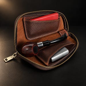 以下が含まれることがあります： ジッパー付きの茶色のレザー製パイプポーチ。中には黒いマウスピースが付いたダークウッドのパイプ、銀色の金属製ライター、内ポケットに収められた小さな赤い長方形のオブジェクトが入っています。