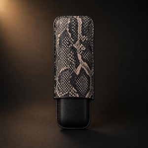 May include: A rectangular cigar case featuring a two-tone design. The upper portion displays a textured black and beige snakeskin pattern, while the lower section consists of a smooth, solid black base. The case has rounded corners and edges.