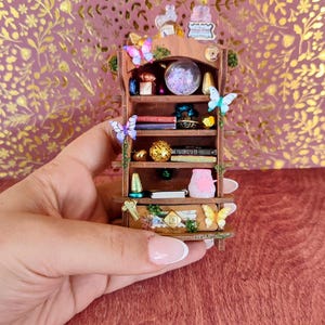 Puede incluir: Una estantería de madera en miniatura llena de libros pequeños, una bola de cristal y objetos decorativos. Los estantes están adornados con mariposas de colores. La unidad se sostiene en una mano contra un fondo floral rosa y dorado.