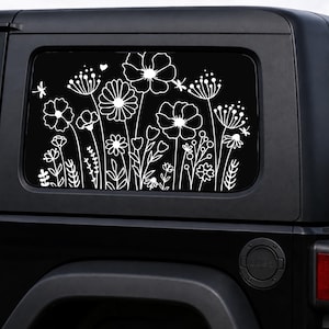 May include: A black vehicle window with a white floral decal. The decal features various stylized flowers, leaves, and a few small insects. The design is a silhouette of wildflowers.