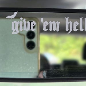 May include: A white vinyl decal applied to a rearview mirror featuring the text "give 'em hell" in a gothic-style font, accompanied by a small silhouette of a bat positioned above the letter g. The decal is applied to the glass surface.