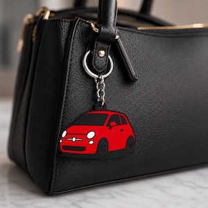 May include: A red car keyring with black detailing hangs from a black leather handbag. The keyring depicts a small car with headlights, windows, and wheels. The handbag has a top handle and a zip closure.