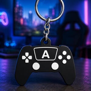 May include: A black keychain shaped like a video game controller with a white D-pad, four white circular buttons, and a central white rectangular accent featuring the letter A in black. The controller is attached to a silver metal ring.