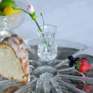 May include: A clear glass serving platter with a radial pattern holds a slice of glazed pound cake, a single fresh strawberry, several blueberries, and a small, faceted glass bud vase containing a single pink and white carnation flower.