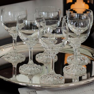 Pode incluir: Um conjunto de taças de vinho de cristal transparente com bases texturizadas e hastes delicadas. As taças estão dispostas numa bandeja prateada, refletindo a luz. As taças estão cheias de um líquido transparente, criando um ambiente elegante.