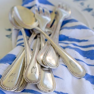 May include: A collection of silver-toned metal cutlery, including spoons and forks, resting on a white cloth with blue horizontal brushstroke patterns. Each piece of flatware features a beaded border along the handle and a small engraved emblem.