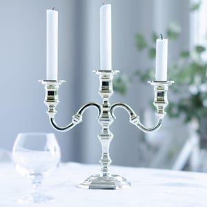 May include: A silver candelabra with three white candles. The candelabra has a decorative, ornate design and is placed on a white surface. A wine glass is in the foreground. The candles are approximately 8 inches tall.