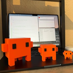 May include: Three orange, blocky figures with black square eyes and legs sit on a laptop. The figures vary in size, with the largest in the foreground. A laptop displays code on the screen, and a white wireless earbud case rests on the laptop.