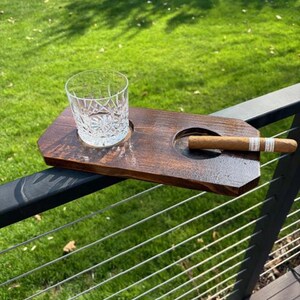 May include: A dark brown wooden cigar and drink holder with a cut crystal glass and a cigar. The holder has a circular indentation for the glass and a larger one for the cigar. The background is green grass.