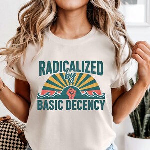 May include: Cream-colored t-shirt with the text "RADICALIZED by BASIC DECENCY" in teal and orange. The design includes a rising sun, waves, and a raised fist. The shirt is a casual wear item.