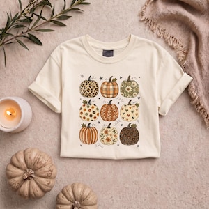 May include: A cream-colored short-sleeve t-shirt featuring a graphic print of nine pumpkins arranged in a grid. Each pumpkin displays a unique pattern, including leopard print, plaid, floral motifs, polka dots, and heart shapes in earthy tones.