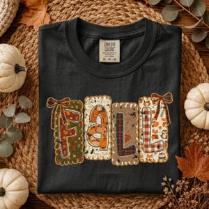 May include: A black short-sleeved t-shirt with a graphic print that spells the word FALL. Each letter is framed in a scalloped border with distinct patterns including pumpkins, plaid, and woodland animals, accented by decorative bows.