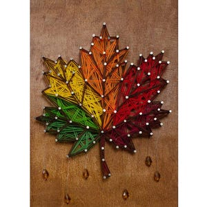 May include: A string art maple leaf design mounted on a brown wooden board. The leaf is created with colourful threads in shades of green, yellow, orange, red, and dark maroon, secured by silver nails. Four small amber-coloured beads hang below.