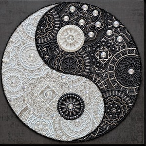 Könnte beinhalten: Ein kreisförmiges Wandkunstwerk mit einem Yin-Yang-Design aus komplizierten Perlenarbeiten und Kristallen. Die linke Seite ist weiß mit silbernen Akzenten, während die rechte Seite schwarz mit dunklen Perlen ist, beide mit geometrischen Mustern.