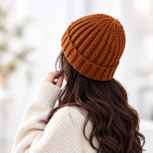 May include: A rust-coloured knit beanie with a thick, vertical ribbed texture and a wide, folded cuff. The hat is constructed from a chunky yarn, featuring a consistent pattern that runs from the crown down to the base of the folded brim.